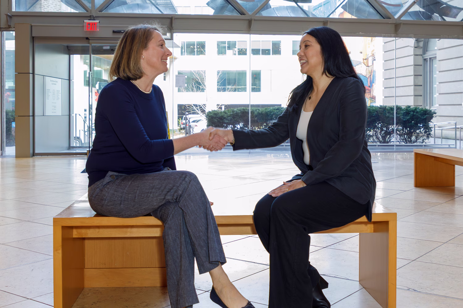 Two businesswomen shaking hands while seated in a modern office lobby